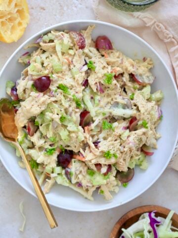 bowl of chicken salad in white bowl with grapes and gold spoon.